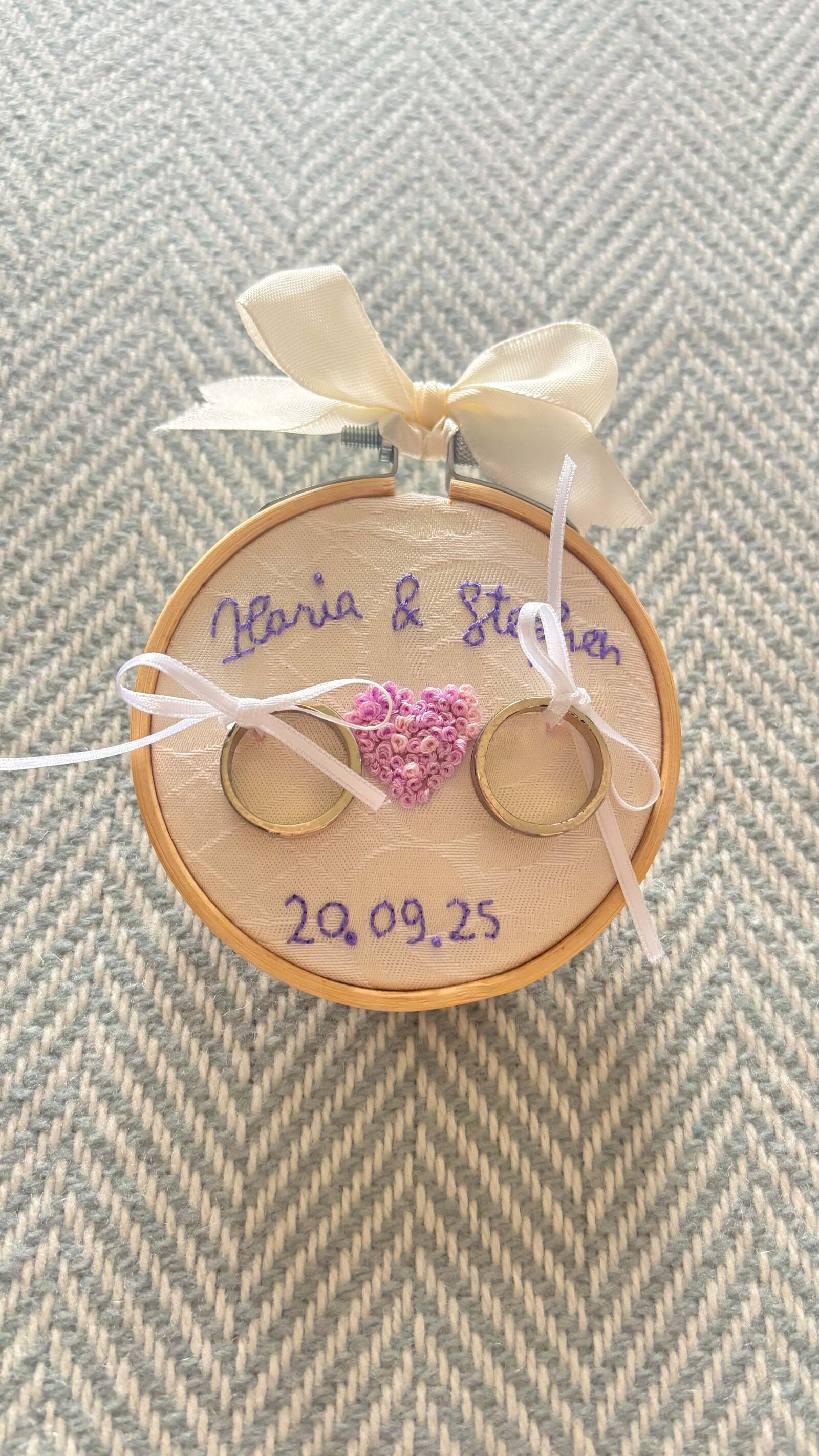 Embroidery hoop ring holder stitched with a couple's names and wedding date, with two rings tied on with white ribbons